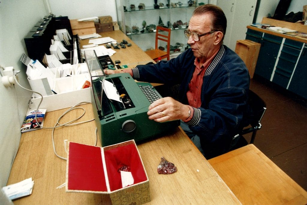 Nissinen skriver vid arbetsbordet med skrivmaskin, bredvid honom på bordet finns en dekorativ låda som innehåller mineralprover eventuellt från Kina.