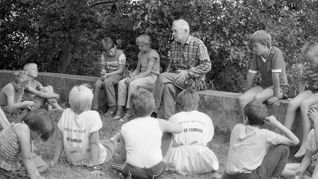 Salkio ja neljä lasta istuvat kivimuurilla. Joukko muita lapsia istuu heidän edessään nurmikolla.