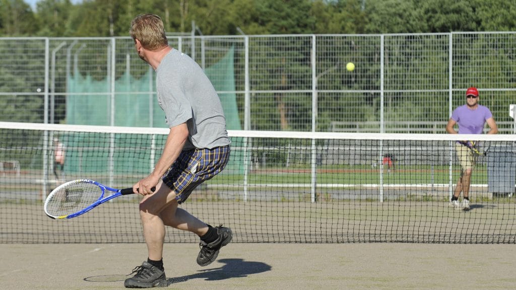 Två män spelar tennis på en utomhusplan. Mannen som är närmare står med ryggen mot kameran och har en blå racket i handen. På sig har han rutiga shorts och en grå t-skjorta. Mannen som står längre bort på andra sidan nätet har på sig en lila t-skjorta, skärmmössa och solglasögon. En gul tennisboll flyger ovanför nätet.