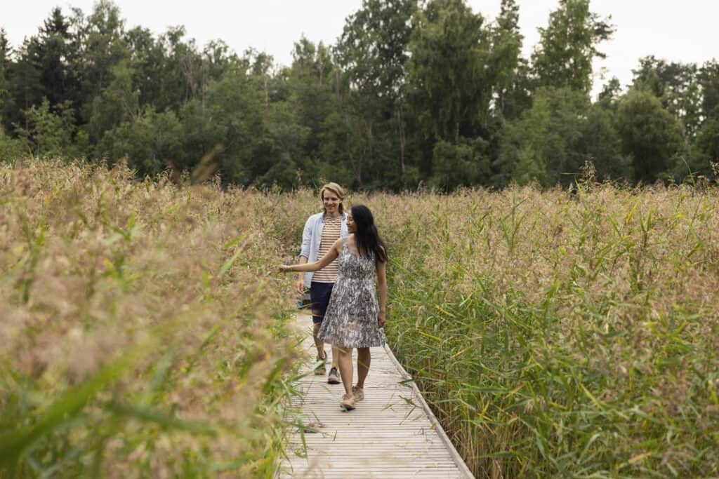 En ung kvinna och en man går efter varandra på träplattformen mitt i vassen. En skog kan ses i bakgrunden. Kvinnan sträcker ut handen mot vassen.