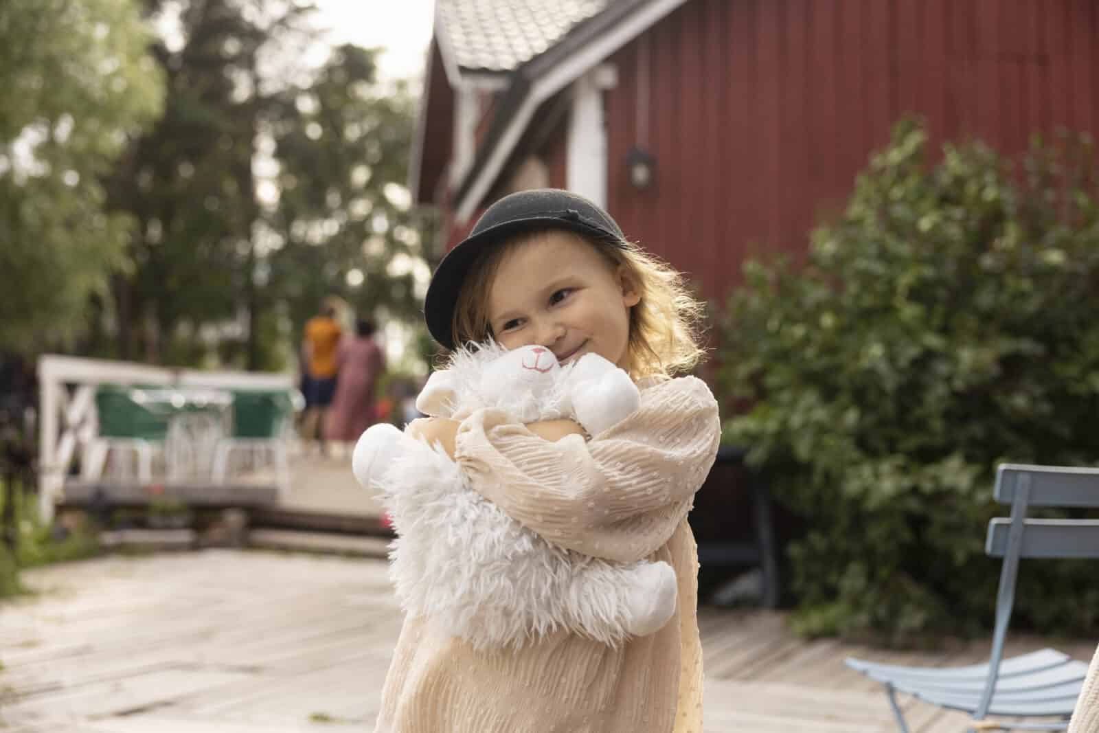 Pieni hattupäinen tyttö rutistaa valkoista pehmolelua ja hymyilee. Taustalla näkyy ihmisiä puisella terassilla ja vihreitä pensaita.