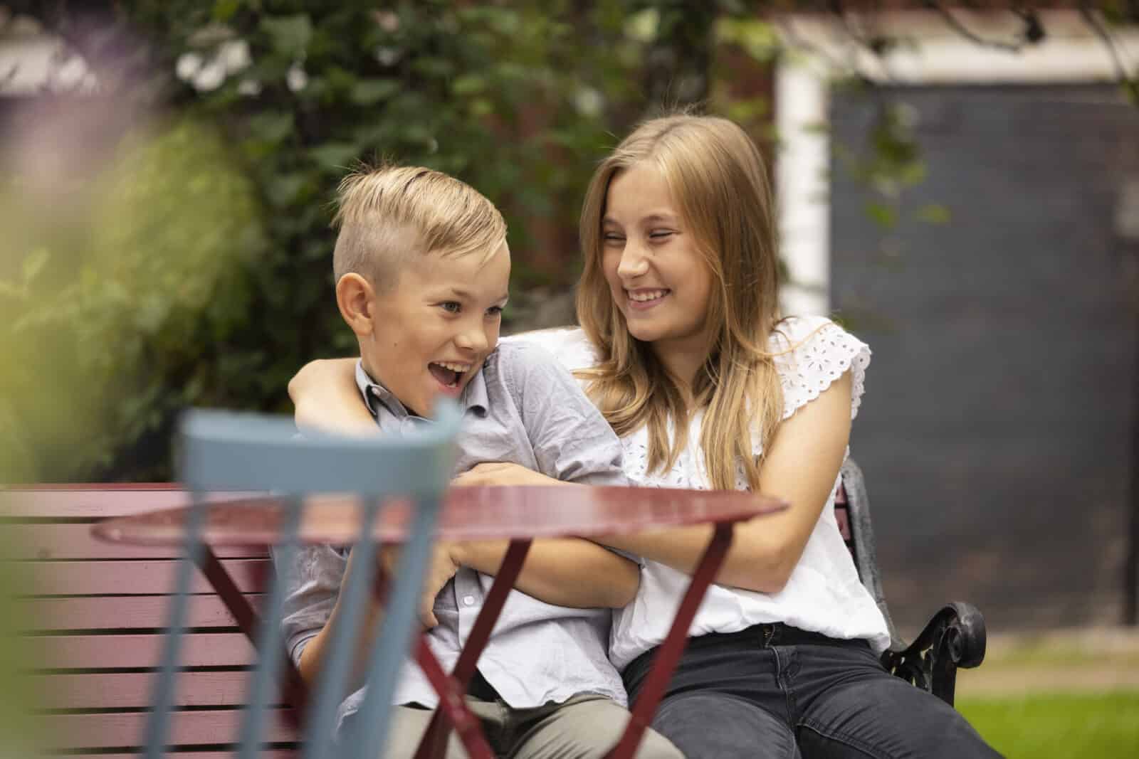En flicka och en pojke sitter i en trädgård. De skrattar tillsammans. I förgrunden kan du se en trädgårdsstol, bakom dörren till gårdsbyggnaden.