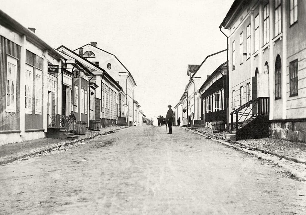 Gammal svartvit bild av Storgatan norrut från korsningen med Fabriksgatan. I mitten står en man med promenadkäpp, iklädd hatt.