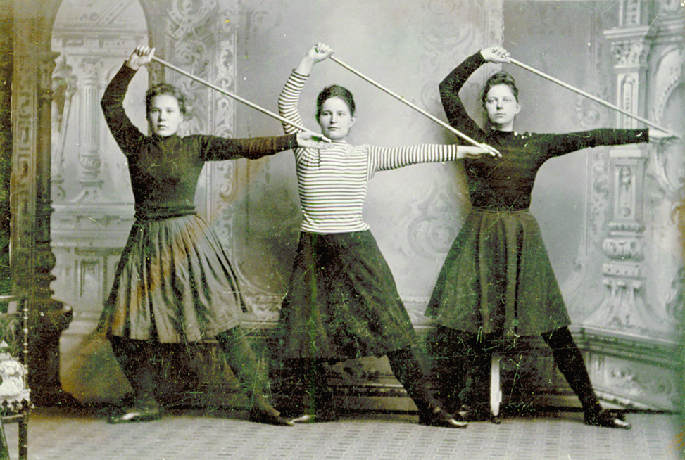 Vanha mustavalkoinen kuva, jossa kolme nuorta naista voimistelee kepin kanssa.