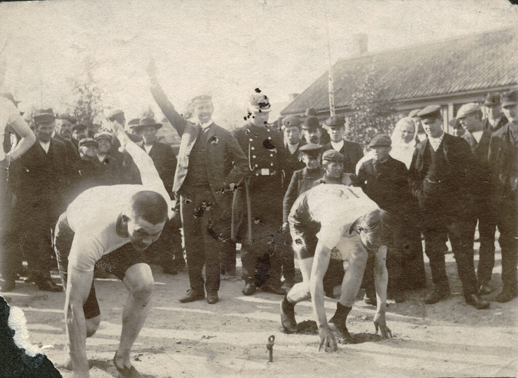 Kuva vuodelta 1907 esittää juoksukilpailun starttitilannetta. Kaksi urheilijaa odottavat starttipistoolin laukaisua yleisön ympäröimänä.