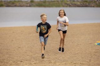 Kouluikäiset tyttö ja poika juoksevat rantahiekalla shortseihin ja t-paitaan pukeutuneena. Pojalla on kädessään frisbee. Meri näkyy taustalla