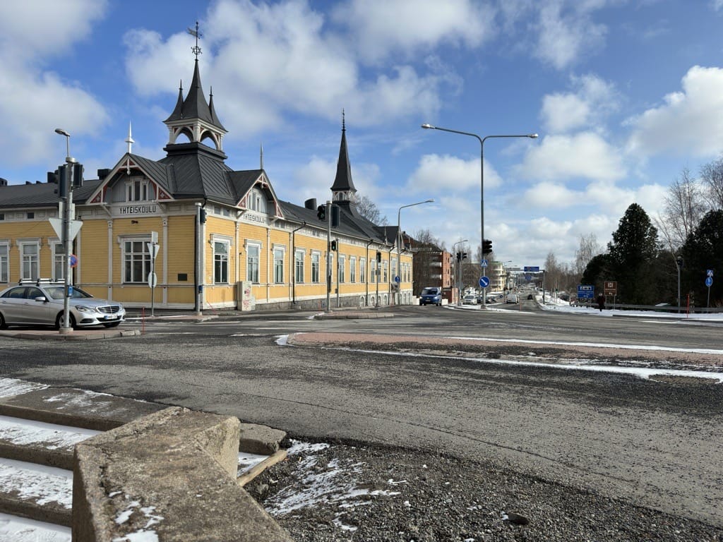 Kevättalvinen katunäkymä. Aurinko paistaa, asvaltilla näkyy hiekotushiekkaa. Taustalla näkyy keltainen puurakennus, jonka kulmissa on koristeelliset tornit.